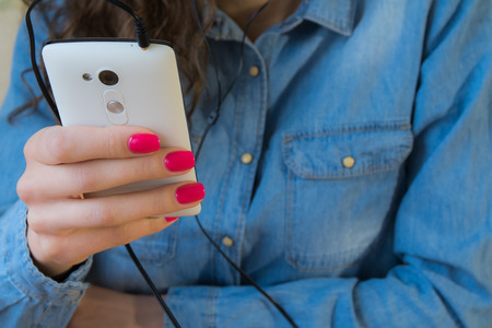 The girl in blue denim shirt with red nail Polish listens to music on a white phone.