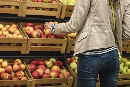 Girl in the beige jacket and blue jeans picks apples in the supermarket. Girl seen from the back. In the background you can see the boxes of apples.の写真素材