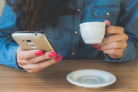 Young girl in a denim shirt sitting in the cafe at the wooden table. In her hands with a red manicure white mobile phone and a cup of espresso. On the table is a white saucer. Vintage photo.の写真素材