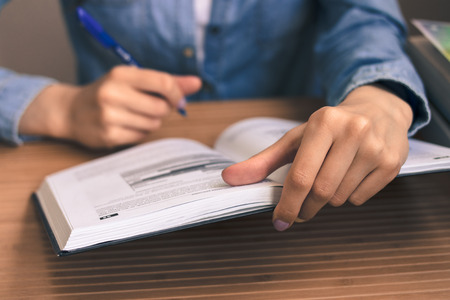 The girl student in a blue denim shirt sitting at a wooden table, reading a textbook and taking...