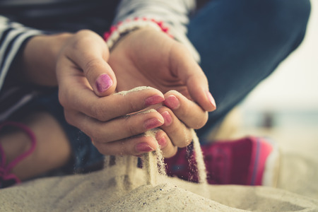 Girl sitting on sand at the beach. In her open palms sand that pours through her fingers. The girl dressed in jeans and pink sneakers, on her arms bracelets and a red manicure on nails. Vintage photo.の写真素材