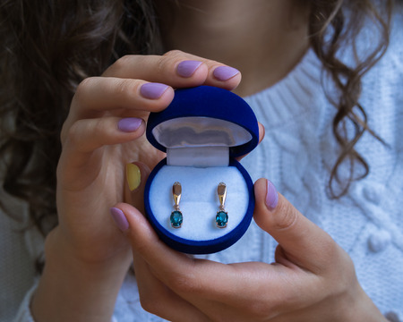 Female hands holding a box of gold earrings with blue Topaz. The girl dressed in a white sweater, her fingers bright manicure. Closeup.の写真素材