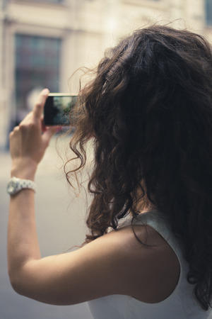 A young girl taking pictures on a smart phone. The girl dressed in a white sleeveless dress. She has curly brown hair. Girl photographs the streets of the tourist town. Closeup photo.の写真素材