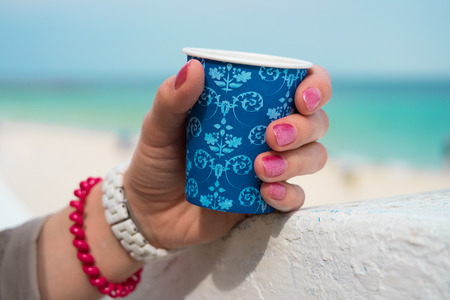 Blue cup of coffee in a female hand on a beach background. The hand rests on a white parapet. On the wrist white and red bracelets.の写真素材