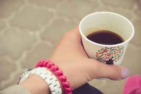 Takeaway coffee in a female hand. The girl is sitting on a park bench and drink coffee. Retro toning.の写真素材