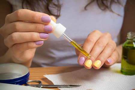 Female hand holding a pipette with oil. A young girl sits at a table and gets a yellow oil on the nails. On the table are nail clippers a bottle of oil a jar of cream and other nail accessories.の写真素材