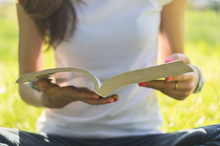 Young woman sitting on a meadow and holding an open book. She is dressed in jeans and a white tshirt. On her hand a ring a watch and a red manicure.の写真素材