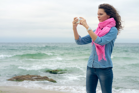 Young woman with curly hair wearing a denim shirt makes the picture of the seaside. Walk along the beach with a camera phone.の写真素材