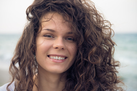 Portrait of young happy smiling woman on the coast in the summer. A woman with brown curly hair resting day on the beach in windy weather. Close up picture.の写真素材