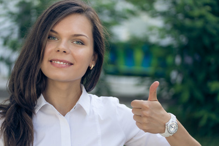 Portrait of a young successful woman in a white shirt with thumb up on a green background outdoors. Smiling pretty girl.の写真素材
