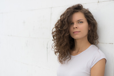 Young beautiful woman with brown curly hair stands near the wall of white brick and looking at the camera. Portrait of a young girl in a white shirt outdoors.の写真素材