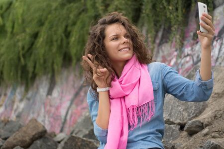 Young happy beautiful smiling woman making a self portrait with the phone camera. Woman sitting on the rocky stones in the green plants. She is wearing a denim jacket and pink scarf.の写真素材