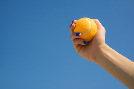 Female hand holds a bright orange tennis ball on a background of blue skyの写真素材