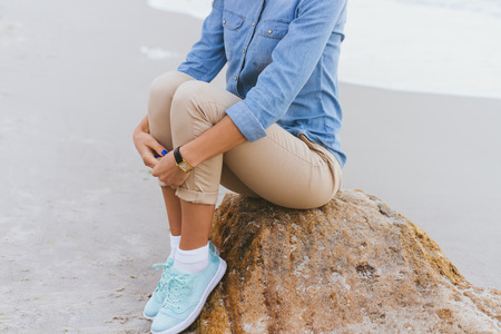 Beautiful young woman sitting on a rock by the sea. She is dressed in beige trousers and a denim shirt. Walking in the fresh air.の写真素材