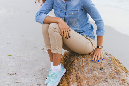 Woman in beige pants and a denim shirt and turquoise sneakers sitting on a rock by the sea. Shirt sleeves rolled up, watch on her arm, a blue manicure.の写真素材