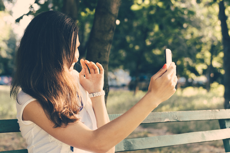 Young woman sitting on a park bench and photographed on a mobile phone. Leisure at outdoors on a sunny day in the summer. Retro colors.の写真素材