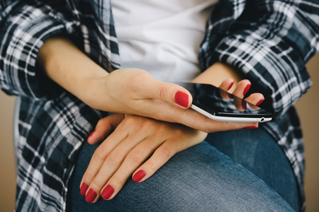 Woman in plaid shirt, jeans and red manicure sitting on a chair and holding a white mobile phoneの写真素材