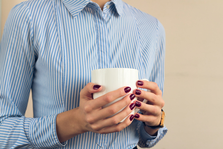 Woman in a blue shirt and red manicure holding a white cup with two hands close upの写真素材