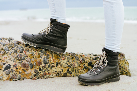 Female feet in blue jeans and black winter boots standing on a stone against the seaの写真素材