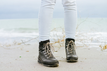 Female feet in blue jeans and black winter boots standing on the beach