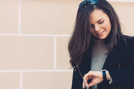 Pretty smiling woman with long hair in black coat checks the time outdoor. Warm toning.の写真素材