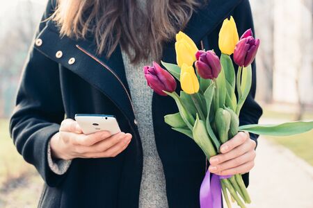 Young woman in a coat holding a bouquet of tulips in one hand and in the other hand mobile phoneの写真素材