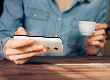 Woman in a denim shirt reading news on mobile phone and drinking coffeeの写真素材