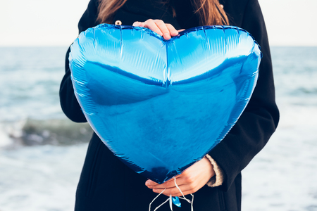 Girl in black coat holding a blue balloon in the shape of a heart on a background of the seaの写真素材