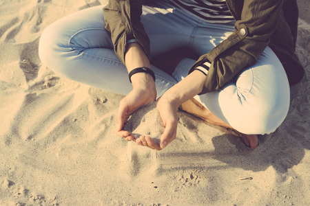 Girl in coat and jeans sitting on the beach cross-legged, in her palms sand. Top view, yellow toning.の写真素材