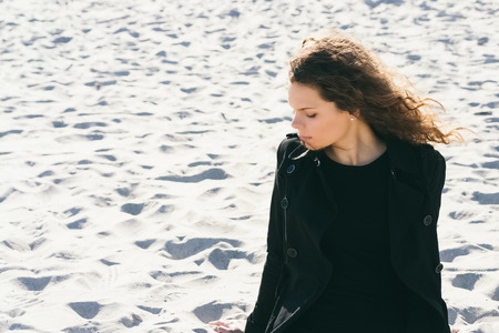 Woman with curly brown hair in a black coat sitting on a pensive at the beach on a sunny day, space for text. Girl sitting on the sand with closed eyes.の写真素材