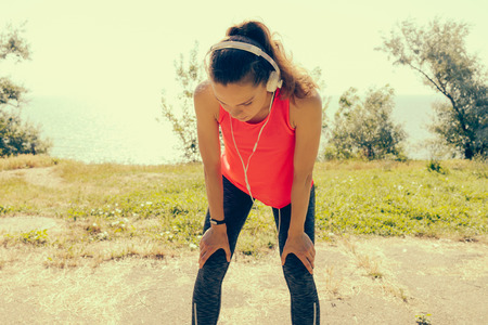 Athletic Girl in headphones and a red T-shirt recovers breath after jogging. Young sporty woman resting after running. Yellow toning.の写真素材
