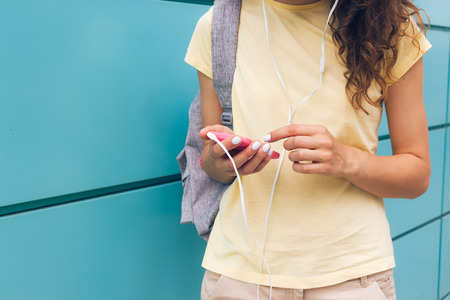 Close-up of female hands with white manicure holding pink mobile phone and headphones outdoors near a blue wallの写真素材