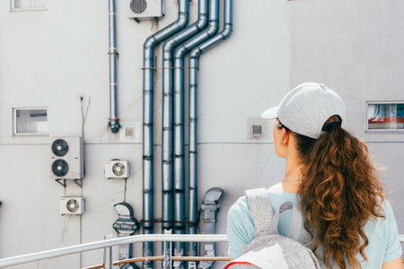 Young woman looks at the wall of the building with air conditioning and ventilation pipesの写真素材