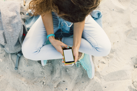 Young slim brunette sits on the beach in jeans and listening to music with headphones and mobile phone, top viewの写真素材