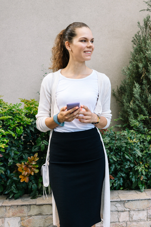 Young woman in a black skirt and white cardigan holding a mobile phone and smiling, vertical frameの写真素材