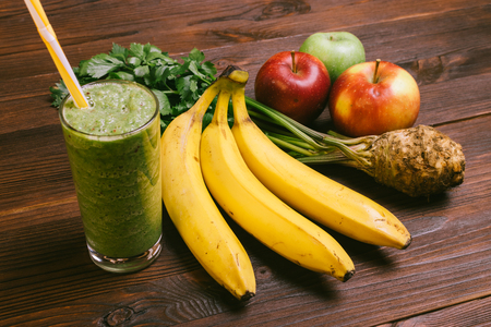 Green smoothie in a glass with a straw, bananas, celery root and apples on a dark wooden tableの写真素材