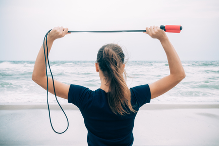 Sport brunette girl standing on the beach and holding a skipping rope over her head, view from the backの写真素材