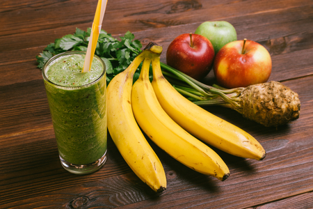Glass of green smoothie with yellow straw on a wooden table next to bananas, apples and celeryの写真素材