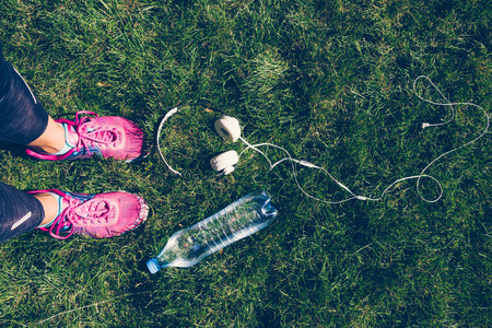 Female legs in pink sneakers, white headphones and a plastic bottle of water on the green grassの写真素材