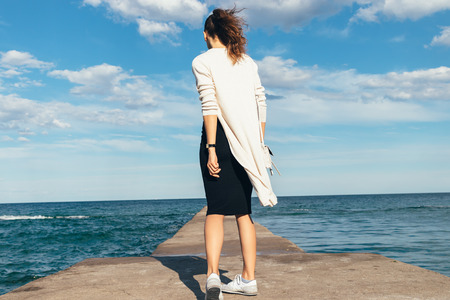 Slim brunette woman in a skirt, cardigan and sneakers walking on the beach in summer, view from the backの写真素材