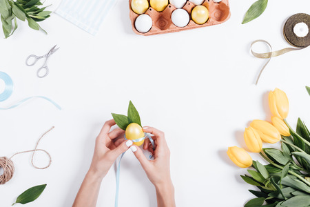 Female hand bandage yellow painted egg blue ribbon. On the table lay a tray with colored eggs, tulips, rope, ribbon and scissors, top view.の写真素材