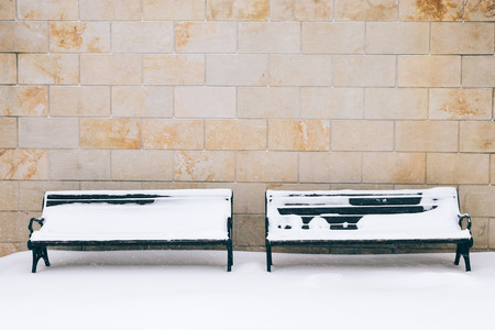 Two snow-covered bench against a brick wall in the winter