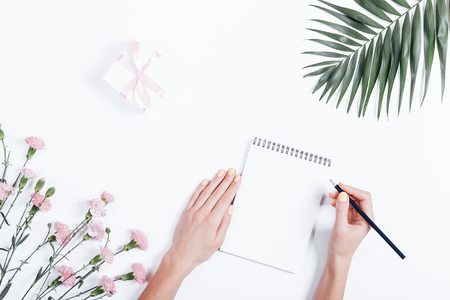 Woman's hand writing in a notebook at the desk, top view. On the white table lay flowers and a box with a gift and ribbon.の写真素材