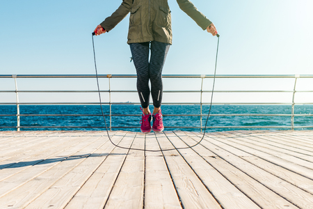 Athletic woman in a jacket and sneakers skipping rope in the morning on the beach, low angle shootingの写真素材