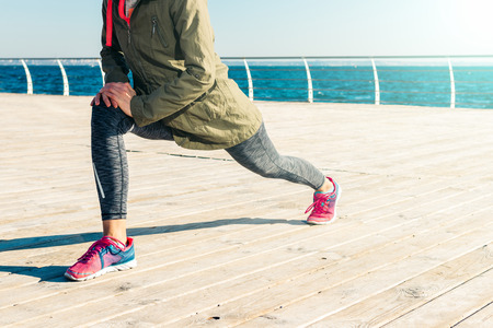 Woman in jacket, hood and sneakers doing stretching on the beach in the morning
