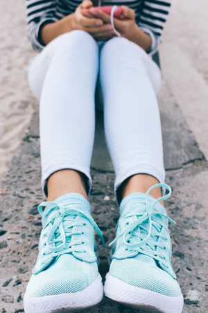 Vertical framing of female legs in jeans and sneakers. Girl in a striped T-shirt listening to music on the beach.の写真素材