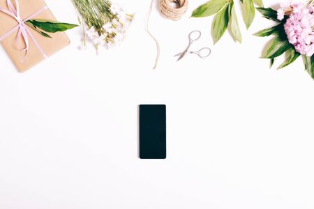 Mobile phone, flowers carnations, gifts, ribbons and packing paper on a white table. Festive composition, top view.の写真素材