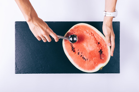 Female hand slices watermelon with a spoon on a white table, top viewの写真素材