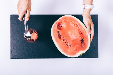 Female hands cut a watermelon with a spoon and put the pieces in a glass on a white table top viewの写真素材