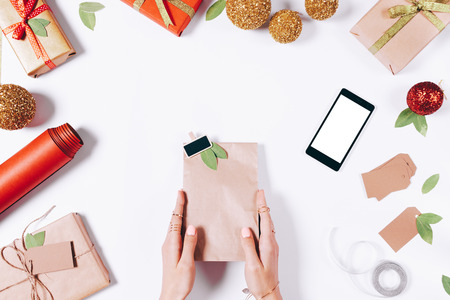 Package with a gift in female hands, a mobile phone and Christmas decorations on a white table top viewの写真素材